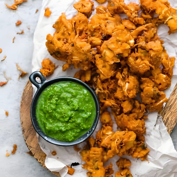 Golden-brown Indian pakoras on white paper with a small black bowl of vibrant green chutney.