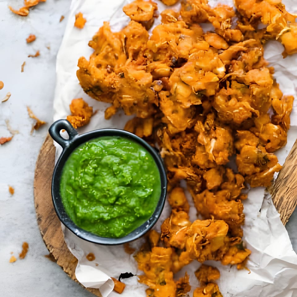 Golden-brown Indian pakoras on white paper with a small black bowl of vibrant green chutney.