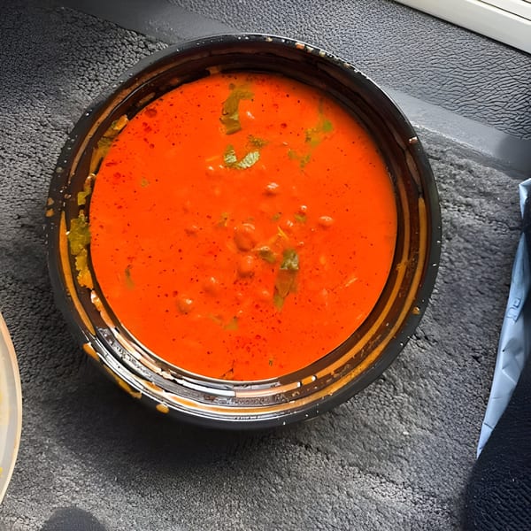 Vibrant orange Indian curry with green herbs in a black takeout container on a textured surface.