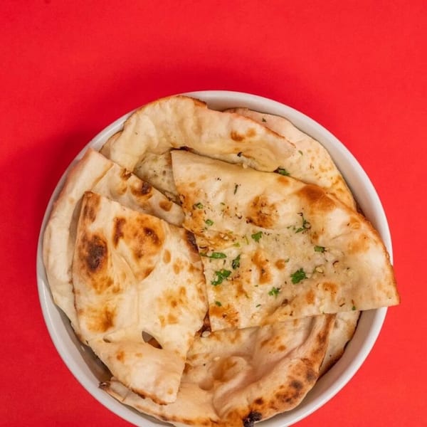 A white bowl filled with golden-brown naan bread, some topped with herbs, against a red background.